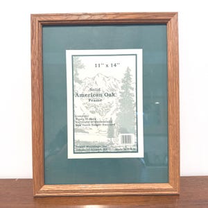 May include: A rectangular solid American oak picture frame with a dark green matting. The center label reads 11" x 14", Solid American Oak Frame, Complete Ready to Hang Vertically or Horizontally, Saw Tooth Hanger Enclosed, Howell Mouldings, Inc.