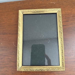May include: A rectangular picture frame with a metallic gold finish and an intricate embossed floral pattern along the border. The frame features a dark, solid-colored insert and a clear glass front that reflects the surrounding environment.