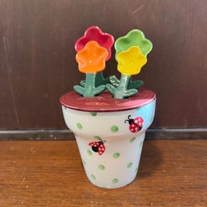 以下が含まれることがあります： 緑色の水玉模様とテントウムシのイラストが描かれた、植木鉢型の白い陶器製計量スプーンセット。赤、オレンジ、黄色、緑のカラフルな花形の持ち手が付いた4本のスプーンが、植木鉢の中に収められています。