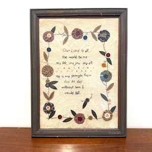 May include: A framed textile art piece featuring embroidered text that reads, "Our Lord is all the world to me my life, my joy, my all. He is my strength from day to day without him I would fall." The border is decorated with fabric flowers and buttons.
