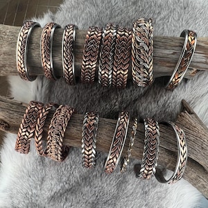 May include: A collection of copper and silver-toned metal bracelets with intricate woven and leaf-like designs. The bracelets are displayed on a piece of driftwood against a backdrop of faux fur. The bracelets vary in width and design.