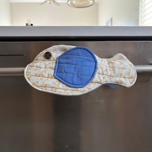 Pode incluir: Uma capa acolchoada em forma de peixe para a pega de eletrodomésticos de cozinha. A capa apresenta um olho azul e um padrão floral em creme e amarelo. Um botão castanho prende a parte superior. Concebido para proteger as mãos de aparelhos quentes.
