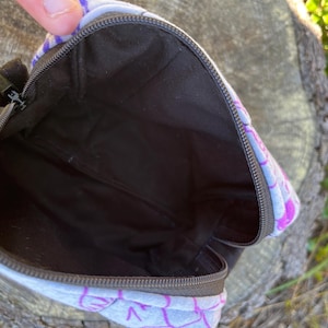 Puede incluir: Bolsa abierta con un dise&ntilde;o estampado. La bolsa tiene un interior negro y un cierre de cremallera. El estampado presenta formas moradas y blancas. Se ve el interior negro.