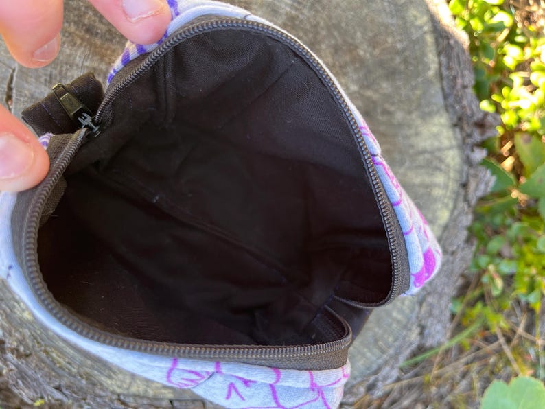 Puede incluir: Bolsa abierta con un dise&ntilde;o estampado. La bolsa tiene un interior negro y un cierre de cremallera. El estampado presenta formas moradas y blancas. Se ve el interior negro.