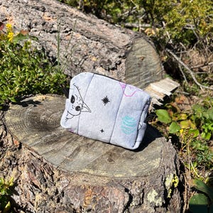 Puede incluir: Un peque&ntilde;o neceser acolchado gris con un personaje de dibujos animados, estrellas y otros dise&ntilde;os. La bolsa es rectangular con esquinas redondeadas y descansa sobre un toc&oacute;n de &aacute;rbol en un entorno natural.