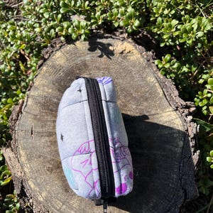 Puede incluir: Una bolsa acolchada gris con cremallera negra, con un dise&ntilde;o caprichoso en morado, verde azulado y blanco. La bolsa descansa sobre un toc&oacute;n de &aacute;rbol desgastado, rodeado de follaje verde.