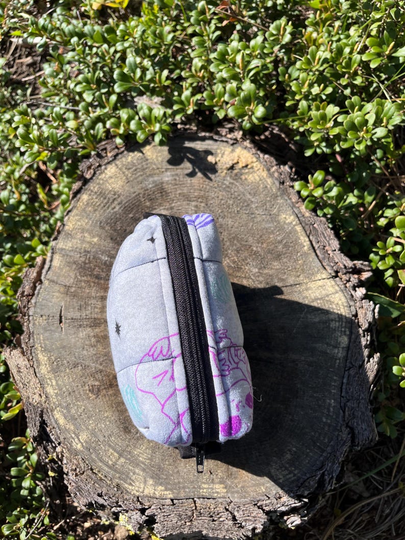 Puede incluir: Una bolsa acolchada gris con cremallera negra, con un dise&ntilde;o caprichoso en morado, verde azulado y blanco. La bolsa descansa sobre un toc&oacute;n de &aacute;rbol desgastado, rodeado de follaje verde.