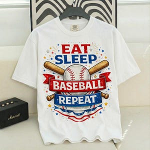 May include: White t-shirt with a baseball-themed graphic. The design includes a baseball, crossed bats, and the words "EAT SLEEP BASEBALL REPEAT" in red and blue. The shirt has a crew neck and short sleeves.