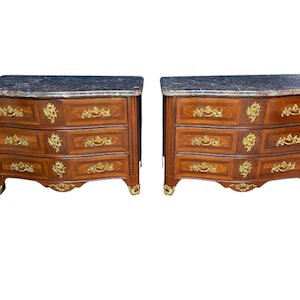 May include: Two matching antique wooden chests of drawers with marble tops. Each chest has three drawers with ornate gold-colored hardware and decorative accents. The wood has a rich, warm brown tone.