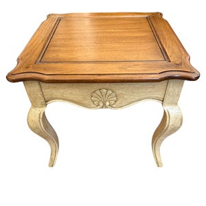 May include: A small, square side table with a light wood top and a darker wood trim. The base is painted a cream color with ornate, curved legs. A decorative shell design is carved into the apron of the table.