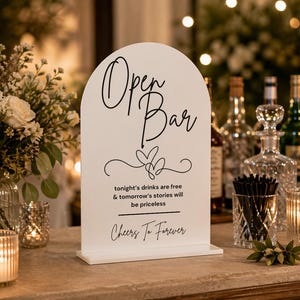 May include: White Open Bar sign with black script. The sign reads "Open Bar" and "tonight's drinks are free & tomorrow's stories will be priceless. Cheers To Forever." The sign is on a table with candles and floral arrangements.