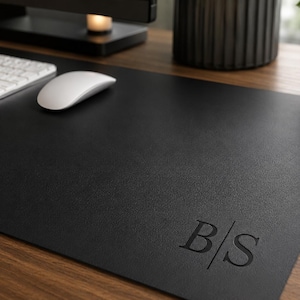 May include: A rectangular black desk mat with a smooth, matte texture. The bottom right corner features a debossed monogram consisting of the capital letters B and S separated by a vertical line. The mat is positioned on a wooden surface.