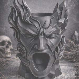 May include: A dark gray decorative planter shaped like a screaming face with stylized flames and crystal-like accents. The planter has an open mouth and closed eyes, with a skull in the background.