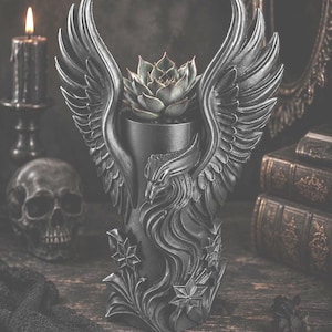 May include: A dark gray, gothic-style planter shaped like a winged creature, holding a succulent plant. The detailed design includes feathers, floral accents, and a textured finish. The background features a skull, candle, and antique books.
