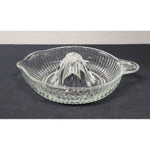 May include: A clear glass citrus juicer featuring a central reamer cone surrounded by a shallow, circular basin with vertical ribbed detailing. The juicer includes a small pouring spout on one side and a sturdy handle on the opposite side.