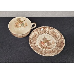 May include: Cream-colored teacup and saucer set with a brown and green pheasant design. The cup has a handle and the saucer has a scalloped edge. The set is likely made of ceramic or porcelain.