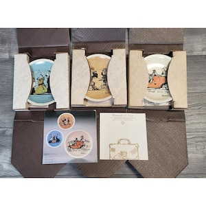 May include: Set of decorative plates featuring vintage illustrations, each in a cardboard display box. The plates showcase scenes with people and vehicles, with text at the bottom. The plates are in a brown box with a brown tie-shaped design.