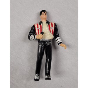 May include: A plastic action figure of a male singer with dark hair, wearing a cream-colored shirt under a black jacket with red and white vertical stripes on the chest. The figure has black pants, white shoes, and is holding a black microphone.