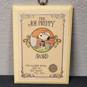 May include: A framed "Joe Preppy Award" featuring Snoopy in a preppy outfit, holding a tennis racket and briefcase. The cream-colored frame has a decorative border and a metal ring for hanging. The text reads "For always being just so, but never too, too!"