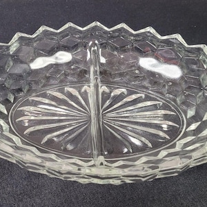 May include: Clear glass divided serving dish with a geometric diamond pattern and a starburst design in the center. The dish has a zig-zag edge and is oval-shaped. Ideal for serving snacks or appetizers.