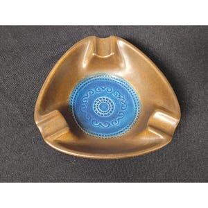 May include: A triangular ceramic ashtray featuring a smooth brown outer rim and a recessed center glazed in a vibrant blue. The blue basin is decorated with a circular pattern of embossed swirls and a central ring of small, repeating dots.