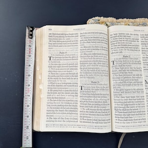 May include: Open Bible with a yellow measuring tape showing the page thickness. The text is from Psalms 18:49 and 19:1. The book is open to a page with text and a white ribbon bookmark.