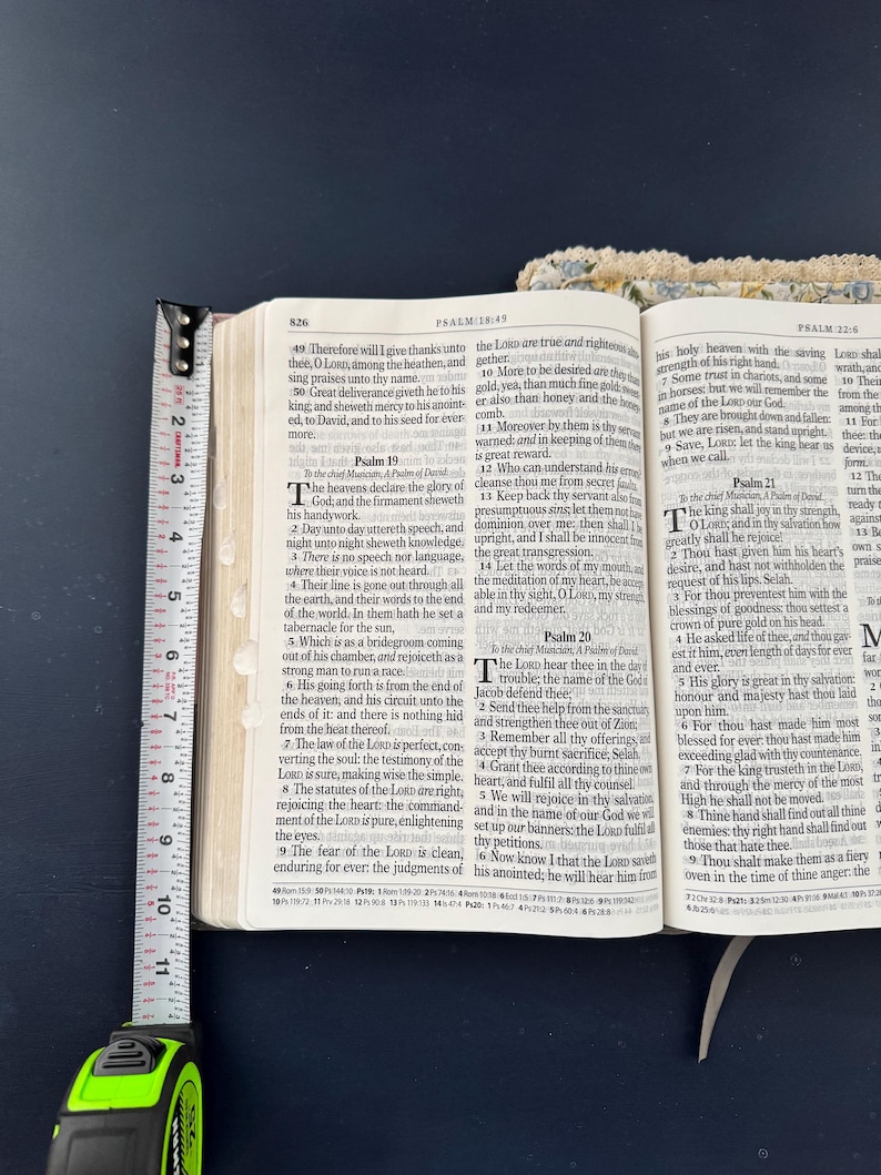 May include: Open Bible with a yellow measuring tape showing the page thickness. The text is from Psalms 18:49 and 19:1. The book is open to a page with text and a white ribbon bookmark.