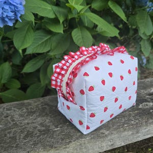 May include: A quilted white pouch featuring a repeating red strawberry pattern and a red and white gingham ruffled trim along the top zipper closure. The rectangular bag has a structured shape with visible diamond-patterned stitching throughout.