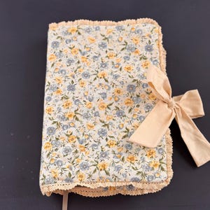 May include: A floral-patterned journal with a cream-colored bow. The journal cover features a light background with yellow and blue flowers. The edges are trimmed with cream-colored lace. A cream-colored ribbon is used as a bookmark.