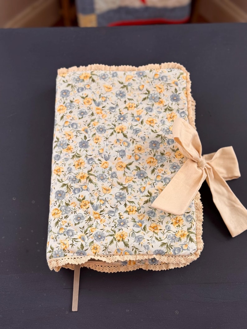 May include: A floral-patterned journal with a cream-colored bow. The journal cover features a light background with yellow and blue flowers. The edges are trimmed with cream-colored lace. A cream-colored ribbon is used as a bookmark.