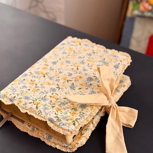 May include: A floral-patterned book cover with a cream-colored lace trim and a matching ribbon tied in a bow. The cover features a pattern of small blue and yellow flowers on a cream background. The book is closed and resting on a dark surface.