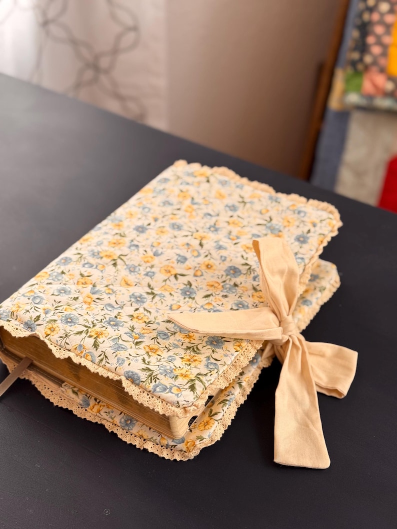 May include: A floral-patterned book cover with a cream-colored lace trim and a matching ribbon tied in a bow. The cover features a pattern of small blue and yellow flowers on a cream background. The book is closed and resting on a dark surface.