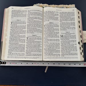 May include: An open Bible with text, resting on a dark blue surface. A measuring tape is placed across the open pages, showing a measurement of approximately 6 inches. The Bible has a light-colored cover with floral accents.