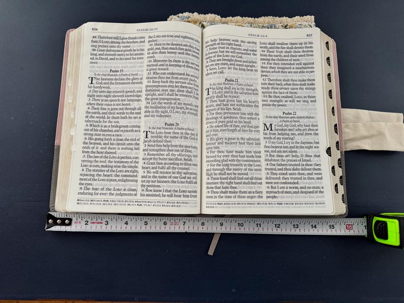 May include: An open Bible with text, resting on a dark blue surface. A measuring tape is placed across the open pages, showing a measurement of approximately 6 inches. The Bible has a light-colored cover with floral accents.