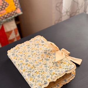 May include: A floral fabric book cover with a cream background and blue and yellow flowers. The cover has a cream-colored lace trim and a cream bow. A cream-colored ribbon bookmark extends from the book.
