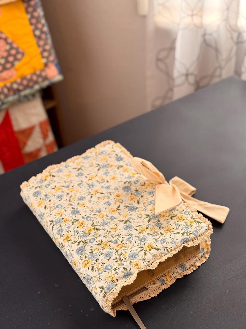 May include: A floral fabric book cover with a cream background and blue and yellow flowers. The cover has a cream-colored lace trim and a cream bow. A cream-colored ribbon bookmark extends from the book.