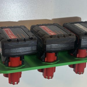May include: A green battery holder with three black and red power tool batteries. The holder is mounted on a white surface. The batteries are arranged in a row, with the red bases visible.