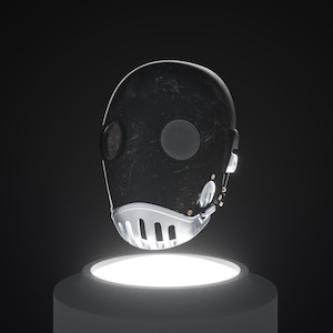Op de afbeelding: Een zwart masker met een futuristisch ontwerp, met ronde openingen voor ogen en mond. Het masker heeft een textuur en wordt getoond op een verlichte, cilindrische sokkel tegen een donkere achtergrond. Het onderste deel van het masker is zilver.