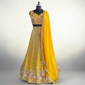 Puede incluir: Un vibrante lehenga amarillo, una prenda tradicional india, exhibido en un maniquí. El conjunto presenta intrincados bordados con patrones florales y geométricos en rojo, blanco y dorado. Un dupatta a juego cae sobre el hombro, completando el look.