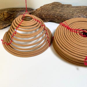 May include: Two tan, spiral-shaped incense coils made of compressed material. One coil is pulled upward into a conical shape, while the other remains flat. Both coils are connected by thin, bright red thread that runs across the surface.