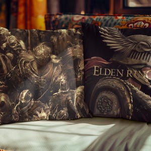 Puede incluir: Dos cojines decorativos con arte de fantasía. Un cojín presenta una ilustración detallada en tonos sepia de figuras con armadura. El otro cojín tiene un fondo negro con un personaje de Elden Ring y el título del juego.