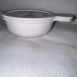 May include: A white bowl with a gray lid and a handle. The lid has text printed on it. The bowl is sitting on a white surface. The bowl is likely used for food preparation or serving.