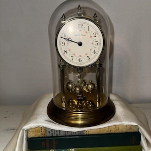May include: A gold-toned anniversary clock with a white face featuring black numerals and a floral pattern, enclosed in a glass dome. The clock has a rotating pendulum with four gold spheres and is marked with the text Black Forest and Made in Germany.