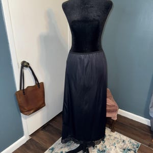 Op de afbeelding: Een zwarte onderrok op een mannequin. De rok is gemaakt van een gladde, glanzende stof en heeft een delicate kanten rand aan de zoom. Een bruine leren tote bag hangt op de achtergrond aan een deurklink.