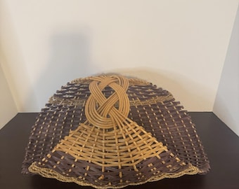 May include: A woven basket featuring a two-tone design with dark brown and light tan reed materials. The basket has a wide, curved shape with a prominent, decorative braided knot detail centered at the top, creating a distinct arched handle structure.