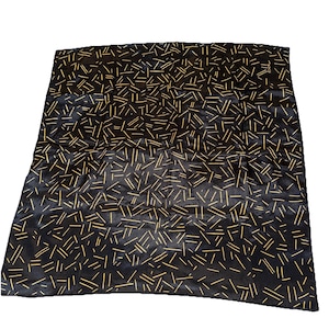 May include: Black square scarf with a repeating pattern of thin, gold-colored lines. The lines are randomly placed across the scarf, creating a textured appearance. The scarf is made of a shiny material.
