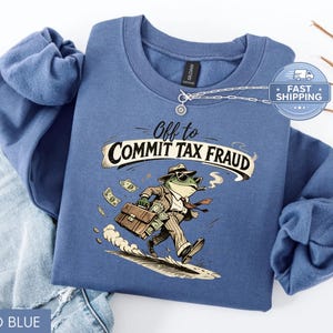 May include: Indigo blue sweatshirt with a humorous graphic of a frog in a suit carrying a briefcase and smoking a cigar, with the text "Off to Commit Tax Fraud". The frog is depicted on a surfboard with money around it.