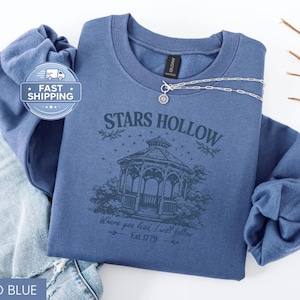 May include: Indigo blue sweatshirt with the text "Stars Hollow" and a gazebo graphic. The text "Where you lead I will follow, Est. 1779" is below the graphic. A silver necklace is draped across the sweatshirt.