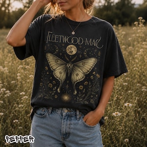 May include: Dark grey t-shirt with a moth graphic and the words "Fleetwood Mac". The moth design includes a full moon and floral details. The shirt has a vintage look. The design is in a gold color.