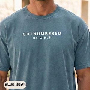 May include: Blue jean colored t-shirt with the text "OUTNUMBERED BY GIRLS" in white block letters. The shirt has short sleeves and a crew neck. The shirt is made of a soft, comfortable material.